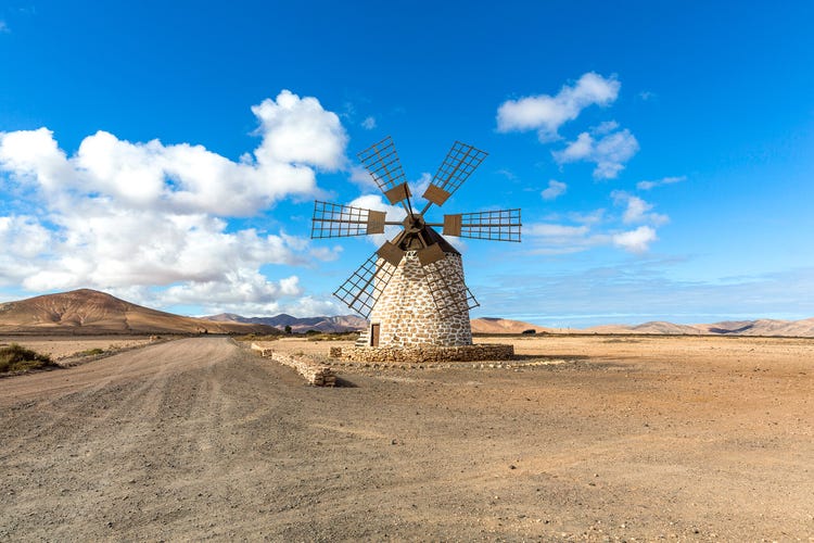 A windmill stands against a blue sky, surrounded by arid landscape and distant mountains in Lajares, Fuerteventura.