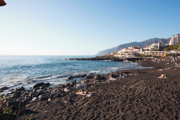 A black sand beach in Playa de la Arena, calm ocean, and visitors relaxing under the sun with cliffs in the background.