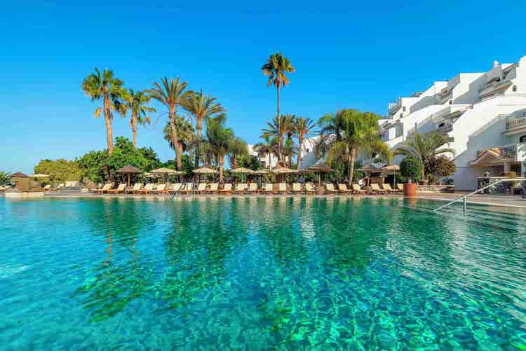 A beautiful pool area in Chayofa, Los Cristianos, surrounded by palm trees, sun loungers, and vibrant greenery.