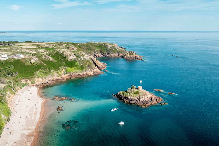 Aerial view of St. Clement, featuring lush green cliffs, a sandy beach, and blue waters with a small tower on a rocky outcrop.
