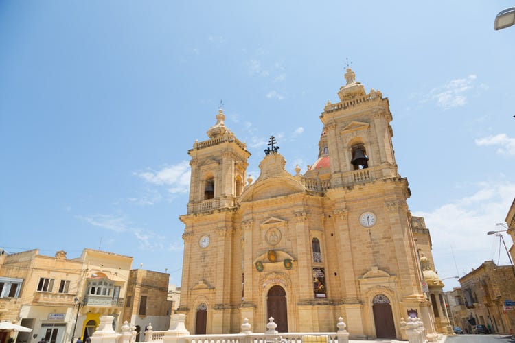 The facade of the Xaghra church in Gozo, featuring two bell towers against a clear blue sky.