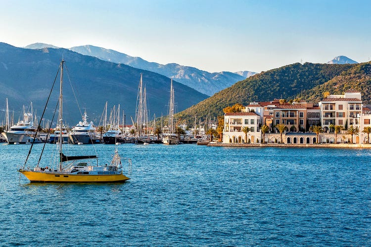 A scenic view of Tivat harbor with luxury yachts and Mediterranean-style buildings against a mountainous backdrop.